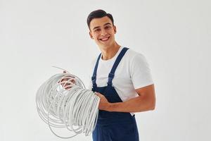 sostiene cable. trabajador de sexo masculino en uniforme azul de pie dentro del estudio contra el fondo blanco foto