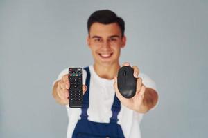 TV remote controller and computer mouse. Male worker in blue uniform standing inside of studio against white background photo