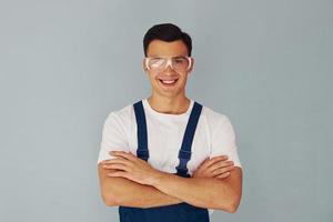 In protective eyewear. Arms crossed. Male worker in blue uniform standing inside of studio against white background photo