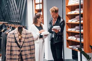 joven trabajadora ayudando a un chico a elegir ropa en una tienda moderna con ropa cara para hombres foto