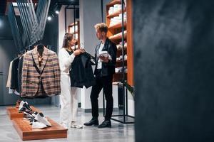 joven trabajadora ayudando a un chico a elegir ropa en una tienda moderna con ropa cara para hombres foto