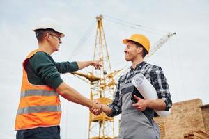 Making handshake. Two construction workers in uniform and safety equipment have job on building together photo