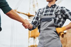 Making handshake. Two construction workers in uniform and safety equipment have job on building together photo