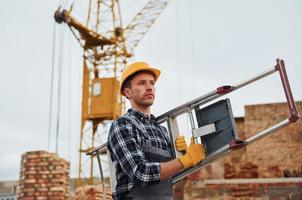 Ladder in hands. Construction worker in uniform and safety equipment have job on building photo