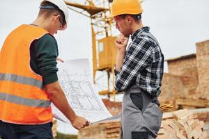 Using paper document with plan on it. Two construction workers in uniform and safety equipment have job on building together photo