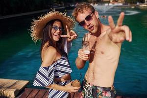 Holding drinks in hands. Cheerful couple or friends together in swimming pool at vacation photo