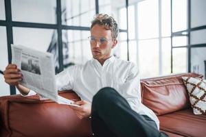 Reads newspaper. Caucasian young guy in elegant white shirt indoors at home photo