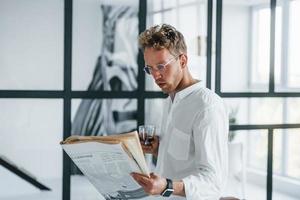 Reads newspaper. Caucasian young guy in elegant white shirt indoors at home photo