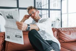 Reads newspaper. Caucasian young guy in elegant white shirt indoors at home photo