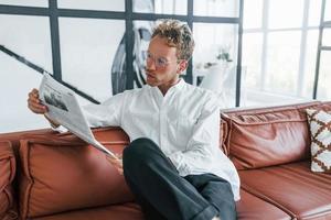 Reads newspaper. Caucasian young guy in elegant white shirt indoors at home photo