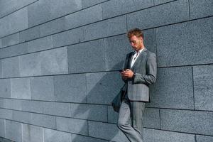 Leans on the wall. Young successful businessman in grey formal wear is outdoors in the city photo