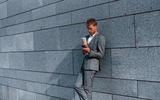 Leans on the wall. Young successful businessman in grey formal wear is outdoors in the city photo