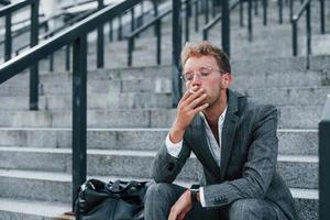 Sits and smokes cigarette. Young successful businessman in grey formal wear is outdoors in the city photo