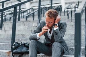 Sits and smokes cigarette. Young successful businessman in grey formal wear is outdoors in the city photo