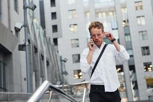 Have conversation by the phone. Young elegant man in good clothes is outdoors in the city at daytime photo