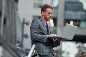 Laptop in hands. Young elegant man in good clothes is outdoors in the city at daytime photo