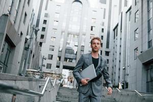 Laptop in hands. Young elegant man in good clothes is outdoors in the city at daytime photo