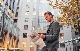 Reads newspaper. Young elegant man in good clothes is outdoors in the city at daytime photo