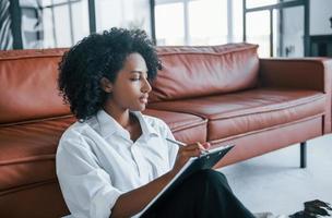 Holds notepad. Young african american woman with curly hair indoors at home photo