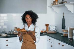 Holds croissants. Young african american woman with curly hair indoors at home photo