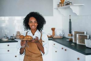 Holds croissants. Young african american woman with curly hair indoors at home photo
