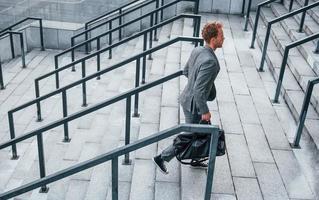 Holds bag in hands. Young businessman in grey formal wear is outdoors in the city photo