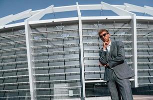Big stadium behind. Young businessman in grey formal wear is outdoors in the city photo