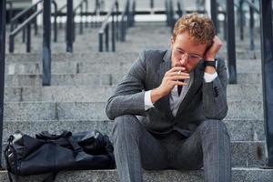 fumando cigarrillos. un joven hombre de negocios con ropa formal gris está al aire libre en la ciudad foto