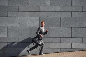 Runs and lating to the job. Young businessman in grey formal wear is outdoors in the city photo