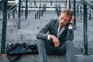 Smoking cigarette. Young businessman in grey formal wear is outdoors in the city photo