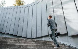 Runs and lating to the job. Young businessman in grey formal wear is outdoors in the city photo