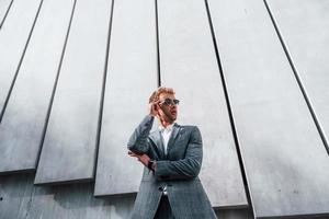 Standing against building wall. Young businessman in grey formal wear is outdoors in the city photo