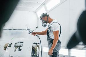 Uses painting gun. Caucasian automobile repairman in uniform works in garage