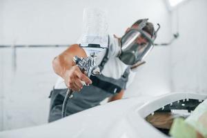 Uses painting gun. Caucasian automobile repairman in uniform works in garage photo