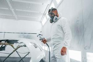 en máscara protectora. pintando el coche. reparador de automóviles caucásico en uniforme trabaja en garaje foto