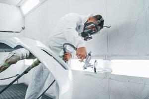 en máscara protectora. pintando el coche. reparador de automóviles caucásico en uniforme trabaja en garaje foto