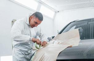 Covering car by brown cloth. Caucasian automobile repairman in uniform works in garage photo