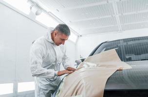 Covering car by brown cloth. Caucasian automobile repairman in uniform works in garage photo