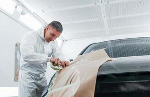 Covering car by brown cloth. Caucasian automobile repairman in uniform works in garage photo