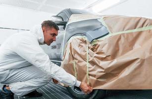 Covering car by brown cloth. Caucasian automobile repairman in uniform works in garage photo