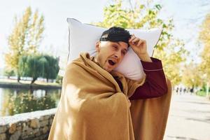 Embracing the pillow. Young man in pajama is outdoors on the street. Feels sleepy photo