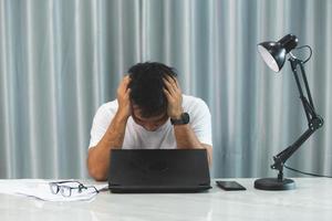 el estresante trabajo de oficina finalmente convierte a un joven en un agotamiento. trabajador masculino estresado, cansado y agotado abrumado por problemas sentado en el escritorio con una computadora portátil y tomándose la mano en la cara foto