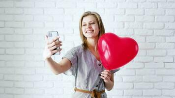 concepto de día de san valentín. hermosa mujer sonriente enamorada charlando y tomando selfie en el teléfono móvil sosteniendo un gran globo de corazón rojo video