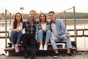 Sitting near lake and embracing each other. Group of young cheerful friends that is outdoors having fun together photo