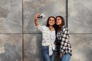 Making selfie. Beautiful cheerful friends or lesbian couple together near wall outdoors at daytime photo