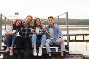 Sitting near lake and embracing each other. Group of young cheerful friends that is outdoors having fun together photo