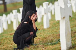 Two young women in black clothes visiting cemetery with many white crosses. Conception of funeral and death photo