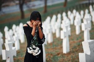 With flowers in hands. Young woman in black clothes visiting cemetery with many white crosses. Conception of funeral and death photo