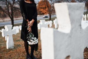 With flowers in hands. Young woman in black clothes visiting cemetery with many white crosses. Conception of funeral and death photo