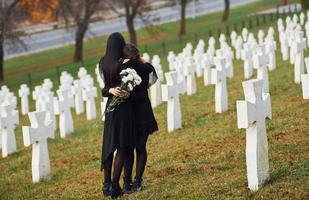 Embracing each other and crying. Two young women in black clothes visiting cemetery with many white crosses. Conception of funeral and death photo